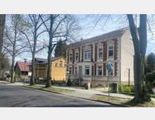 Straßenszene mit einem zweistöckigen Gebäude im klassischen Stil, charakterisiert durch rote Ziegel und weiße Stuckverzierungen. Rechts und links daneben befinden sich kleinere Häuser, während die Umgebung von Bäumen und einem Gehweg gesäumt ist.