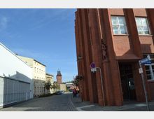 Straßenszene mit Kopfsteinpflaster und Gebäuden, darunter ein rotes Backsteingebäude mit großen Fenstern und einer Skulptur an der Fassade. Im Hintergrund ein Turm mit dunkelgrauem Dach, umgeben von verschiedenen Gebäuden unter einem blauen Himmel.