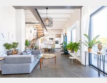 Helles, modernes Wohnzimmer mit offener Küche, großer Fensterfront und Pflanzen. Eine Metall-Holz-Treppe führt in ein oberes Stockwerk, und ein graues Sofa sowie ein runder Glastisch prägen den Wohnbereich.