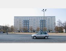 Ein sechsstöckiges Wohngebäude mit heller Fassade und blauen Balkonen, umgeben von kahlen Bäumen entlang eines Gehwegs. Im Vordergrund ein geparkter silberner Kleinwagen auf einer mehrspurigen Straße.