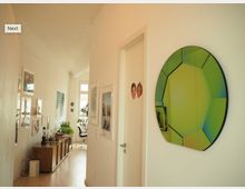 Ein hell beleuchteter Flur in einer Wohnung mit weiß gestrichenen Wänden, dekoriert mit modernen Bildern und einem großen, grünlich spiegelnden Wandobjekt. Im Hintergrund ist ein Wohnzimmer erkennbar, mit Holzmöbeln, Pflanzen und großen Fenstern, die Tageslicht hereinlassen.