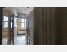 Blick in einen alten, verfallenen Raum mit freistehender Holztür, gelblichen Wandfliesen und einem großen, mehrteiligen Fenster. Im Hintergrund ist ein Heizkörper unterhalb des Fensters zu sehen, die Decke zeigt deutliche Spuren von Schäden.