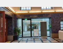 Innenansicht eines öffentlichen Gebäudes mit bläulich gefliesten Wänden, einem Glastürbereich mit der Aufschrift "Kein Durchgang" und einem beleuchteten Notausgangsschild. Im Vordergrund befinden sich eine Topfpflanze und ein aufgestelltes Informationsbrett auf einem schwarz-weißen Fliesenboden.