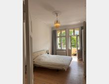 Ein Schlafzimmer mit einem Bett mit weißer Bettwäsche, einem Nachttisch mit Lampe und Holzboden. Große Fenster und eine geöffnete Tür führen zu einem Balkon mit Blick auf grüne Bäume; eine dekorative Deckenlampe hängt in der Mitte des Raumes.