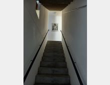 Treppe in einem schmalen, weiß gestrichenen Treppenhaus mit schwarzen Metallgeländern an beiden Seiten; an der Wand oben hängt ein gerahmtes Schwarz-Weiß-Foto, und die Decke ist mit einer einfachen runden Lampe ausgestattet.