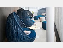 Ein moderner Lounge-Bereich mit blauen, geflochtenen Sitzkapseln, die mit Kissen und Polstern ausgestattet sind. Kleine runde Beistelltische und Hocker in Blau und Weiß ergänzen die Einrichtung, umgeben von einer hellen, minimalistischen Atmosphäre mit Tageslicht.