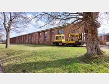 Backsteinhalle mit großen, hohen Fenstern, davor eine gelbe Schienenarbeitsmaschine auf Gleisen. Im Vordergrund ist eine Grasfläche mit Bäumen zu sehen.