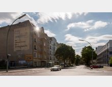 Straßenszene mit Wohngebäuden auf der linken Seite, darunter ein Altbau mit Werbeschrift an der Fassade, und modernen Gebäuden auf der rechten Seite. Bäume säumen die Straße, die von Autos befahren wird, während sich im Hintergrund eine Bushaltestelle befindet.