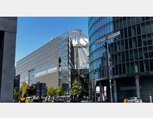 Straßenszene in Berlin mit modernen Gebäuden, darunter der Bahntower mit Glasfassade rechts und ein rechteckiges Bürogebäude links. Im Hintergrund sichtbar das markante Zeltdach des Sony Centers am Potsdamer Platz.