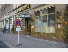 Außenansicht eines Restaurants mit dem Namen „Max und Moritz“ auf einem mit gelben Buchstaben beschrifteten Schild. Die Fassade ist mit grünlich-bräunlichen Keramikfliesen verkleidet, neben dem Eingang befinden sich Laternen, und ein Halteverbotsschild steht auf dem Gehweg.