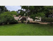 Gartenanlage mit großer Rasenfläche, einem zentral stehenden Baum und umlaufenden Beeten mit Sträuchern. Im Hintergrund verläuft eine hohe Ziegelmauer mit roter Abdeckung, dahinter ragt ein Wohnhausdach hervor; rechts ist ein kleiner Gewächshausbau zu sehen.