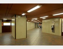 U-Bahn-Unterführung mit orangefarbener Decke und rechteckigen Deckenleuchten, beigefarbenen Wandfliesen und Bodenfliesen in Brauntönen. Im Hintergrund sind Werbeplakate und Personen zu sehen.