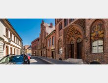 Historische Straßenansicht mit einer gotischen Backsteinfassade auf der rechten Seite, die ein aufwendig verziertes Portal und bunte Wandmalereien zeigt. Links stehen moderne Wohngebäude mit hellem Putz, und parkende Autos säumen die Kopfsteinpflasterstraße.