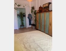 Ein Schlafzimmer mit einem Holzboden, einem beigefarbenen Bettbezug im Vordergrund, einem Vintage-Schrank mit Glastüren und einem alten Radio darauf. Im Hintergrund befinden sich mit Pflanzen dekorierte Wandregale, ein türkis gekachelter Türdurchgang und eine aufgehängte Kleidung.