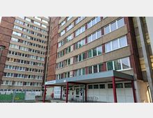 Eingangsbereich eines hohen Wohnblock-Komplexes mit rauer Betonfassade und vielen horizontalen Fensterreihen; ein flaches Vordach auf roten Stützen markiert den Zugang. Links stehen weitere mehrstöckige Gebäudeflügel, im Vordergrund sind Zaun, Garagentore und befestigte Wege zu sehen.