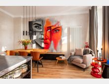 Moderner Wohn- und Essbereich mit Holztisch und Lederstühlen, schwarzer Pendelleuchte sowie einer Sitzecke mit Sessel, kleinem Couchtisch und Beistelltisch. An der Wand hängen großformatige, farbige Porträtgemälde; daneben stehen ein hoher weißer Heizkörper und ein bodentiefes Fenster mit grauen Vorhängen.