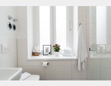 Helles, modernes Badezimmer mit weißen Fliesen und einem großen Fenster, auf dessen Fensterbank dekorative Elemente wie eine Pflanze, Handtücher und Toilettenpapier stehen. Rechts befindet sich eine Duschkabine mit Glaswand, und links ein Waschbecken neben einer Wandhalterung mit weiteren Handtüchern.