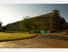 Großes Backsteingebäude mit langen, schmalen Fenstern und einem Satteldach, teilweise mit Baugerüsten versehen. Im Vordergrund eine gepflasterte Straße und eine Grünfläche mit Bäumen bei Sonnenuntergang.