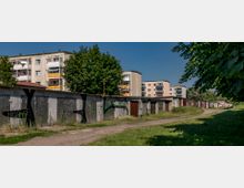 Reihen von flachen Garagen mit bemalten Türen und verwitterten Fassaden, umgeben von Grasflächen und Bäumen, im Hintergrund mehrstöckige Plattenbauwohnungen mit Balkonen.