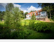 Weißes Landhaus mit rotem Ziegeldach und halbrunden Fenstern, umgeben von einer grünen Wiese, Bäumen und einem kleinen Teich im Vordergrund; das Gebäude steht auf einer leicht erhöhten Natursteinmauer.
