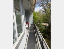 Schmaler Balkon an einem Wohngebäude mit grauem Boden, Metallgeländer und großer Fensterfront; am Ende stehen ein kleiner Tisch und Stühle. Der Balkon blickt auf dichtes Grün mit hohen Bäumen und eine Straße im Hintergrund.
