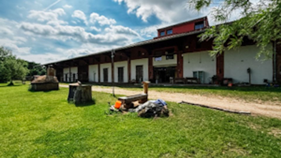 Ländliches Hofgelände mit einem langen, niedrigen Wirtschaftsgebäude mit weißer Fassade und dunklem Dach, davor ein Schotterweg und Grünfläche. Im Vordergrund liegt ein kleiner Haufen Material auf dem Rasen; im Hintergrund sind Bäume und ein wolkiger Himmel zu sehen.