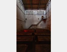 Innenansicht einer verlassenen Industriehalle mit hohen weißen Ziegelwänden, rechteckigen Fenstern in der oberen Wandhälfte und einer Metalltreppe, die zu einer kleinen Plattform führt. Im Vordergrund befinden sich große rechteckige Betonstrukturen, der Raum wirkt abgedunkelt.