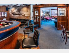 Barbereich mit maritimer Einrichtung, einschließlich einer Theke aus Holz mit Lederhockern und einem großen Wandbild eines Segelbootes. Im Hintergrund ist ein angrenzender Speiseraum mit Holzmöbeln und blauem Teppichboden sichtbar.