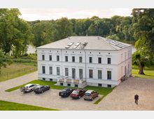 Ein zweistöckiges, weiß verputztes Herrenhaus mit symmetrischer Fassade und großen Fenstern, umgeben von einer Grünfläche und Bäumen, nahe einem See. Vor dem Gebäude befinden sich einige geparkte Autos und ein gepflasterter Vorplatz.