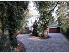 Einfamilienhaus mit heller Fassade, umgeben von hohen Bäumen und dichtem Grün, vor einer geschwungenen gepflasterten Einfahrt mit integriertem Garagengebäude.