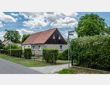 Ein eingeschossiges Wohnhaus mit Satteldach und teilweise holzverkleideter Giebelseite, umgeben von einem Garten und einer Hecke. Im Vordergrund verlaufen ein Gehweg, ein Straßenschild mit den Beschriftungen „Schusterweg“ und „Glaserweg“ sowie eine kleine Straße.