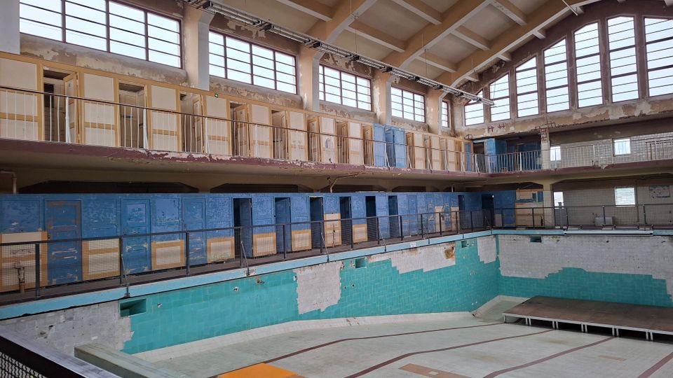 Ein leerstehendes Hallenbad mit abgeblättertem Putz und Farbresten an den Wänden. Die Hauptebene zeigt eine leere Schwimmhalle mit türkisfarbenen Kacheln, umgeben von blauen Umkleidekabinen und einer Empore mit Geländern unter einer hohen Fensterfront.