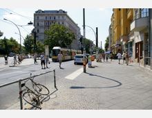 Straßenszene in einer Stadt mit einer gelben Straßenbahn auf den Gleisen, flankiert von historischen und modernen Gebäuden. Fußgänger und Radfahrer bewegen sich auf Bürgersteigen, während ein beschädigtes Fahrrad an einem Geländer befestigt ist.
