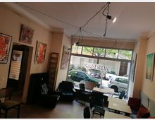 Innenraum eines kleinen Cafés oder einer Galerie mit mehreren Tischen und Stühlen sowie Sesselbereich, beige Wände sind mit großformatigen Gemälden dekoriert. Große Schaufensterfront zur Straße mit Blick auf parkende Autos; an der Decke verlaufen Lichterketten und Spots.