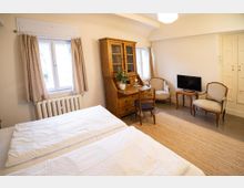 Helles Gästezimmer mit zwei Betten, großen Fenstern mit Vorhängen und Heizkörper darunter. Ein Holzsekretär mit Vitrine, zwei Polstersessel, ein kleiner Tisch mit Fernseher sowie ein heller Teppich auf Fliesenboden ergänzen die Einrichtung.