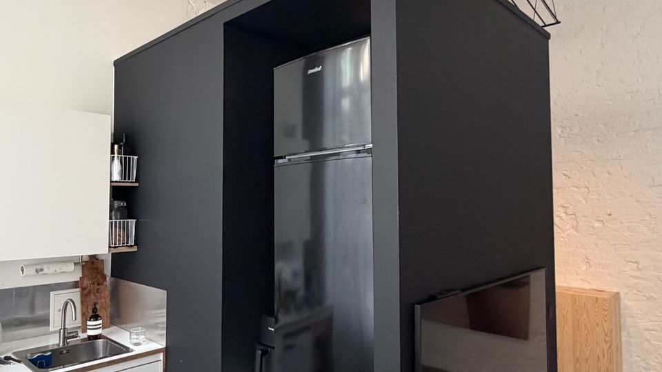 Innenansicht einer modernen Küche in einem Loft mit weiß gestrichenen Deckenbalken und Betonboden. Ein großer, schwarzer Einbaukubus integriert Kühlschrank und Waschmaschine; daneben stehen weiße Küchenschränke mit Spüle und Arbeitsfläche.