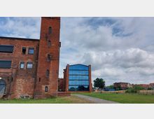 Backsteinbau mit historischer Fassade und kleinem Turm links im Bild, daneben ein modernes Gebäude mit einer Glasfassade. Im Vordergrund befindet sich eine Wiese mit einem gepflasterten Weg, im Hintergrund sind weitere Gebäude und Bäume sichtbar unter bewölktem Himmel.