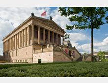 Historisches Gebäude in klassizistischer Architektur mit säulenverzierter Fassade, einer Treppe mit Skulpturen und einer Reiterstatue am Eingang, umgeben von Grünflächen und Bäumen. Der Himmel ist leicht bewölkt, und eine rote Fahne weht auf dem Dach.