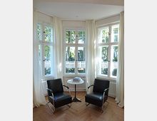 Helle Sitzecke in einem Raum mit drei großen, bodentiefen Fenstern, weißen Vorhängen und einem Fischgrät-Holzboden. Zwei Ledersessel mit einem kleinen runden Tisch in der Mitte, im Hintergrund ist ein weißer Gartenzaun sichtbar.