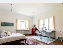 Ein helles Schlafzimmer mit Arbeitsbereich. Links steht ein Bett mit gemusterten Kissen, rechts ein Schreibtisch mit Computer und Bürostuhl vor großen Fenstern mit Vorhängen; der Boden besteht aus Parkett und Teppichen.