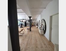 Langer Raum mit Holzboden, weißen Wänden und freiliegenden, weißen Deckenbalken. Links sind Möbel wie ein Schaukelstuhl und ein Boxsack zu sehen, rechts befinden sich Türen und schwarze Wandtafeln mit Notizen.