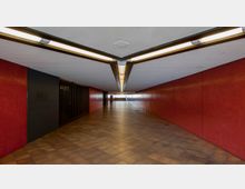 Ein langer, geschlossener Fußgängertunnel mit roten gefliesten Wänden, einem braunen Fliesenboden und einer schräg verlaufenden Decke mit zentraler Leuchtstoffbeleuchtung. Am Ende des Tunnels sind Treppen erkennbar, die ins Freie führen.