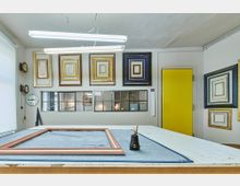 Werkstatt für Bilderrahmen mit einem Arbeitstisch im Vordergrund, auf dem ein Rahmen liegt. An der Wand hängen zahlreiche dekorative Bilderrahmen aus Holz und Gold, daneben befindet sich eine gelbe Tür und ein Fenster mit mehreren kleinen Glasscheiben.