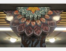 Geometrisch gestaltete Säule in einem Innenraum, verziert mit einem bunten Muster aus stilisierten Blättern in Rosa-, Lila- und Türkistönen. Im Hintergrund sind goldene horizontale Streifen und leuchtende Deckenlampen zu sehen.