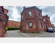 Ziegelbau-Komplex mit historischer Industriearchitektur, bestehend aus mehreren Backsteingebäuden mit Rundbogenfenstern. Auf einem Gebäude sind Schilder mit Beschriftungen wie „Villa Hotel“ und „Speicherhotel“ angebracht, im Vordergrund ein gepflasterter Weg und ein parkendes Auto.