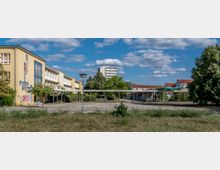 Weitläufiger Außenbereich einer verlassen wirkenden Schule mit einem gelb gestrichenen Hauptgebäude, Graffiti an den Wänden und einer überdachten, teilweise bewachsenen Konstruktion im Hof. Im Hintergrund sind Wohngebäude und Bäume unter einem blauen Himmel mit vereinzelten Wolken zu sehen.