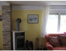 Wohnzimmeransicht mit einer gelben Wand, einem modernen Kaminofen vor einer gemauerten Ziegelwand und einem kleinen Holzlager darunter. Rechts davon ein großes Fenster mit halb geöffnetem weißen Vorhang, daneben ein orangefarbenes Sofa mit gestreiften Kissen.