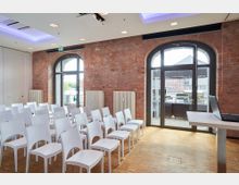 Ein modern eingerichteter Seminarraum mit weißen Stühlen in Reihen auf hellen Holzfußboden. Die Ziegelwände enthalten große Rundbogenfenster, die einen Blick auf eine Terrasse und ein Außengebäude ermöglichen.