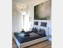 Helles Schlafzimmer mit einem großen Bett mit grauer Bettwäsche, einem weißen Polsterkopfteil und einer Glas-Nachttischlampe. Im Raum befinden sich ein Kronleuchter, ein großer Kleiderschrank mit weißen Türen und ein Bild mit einem Waldmotiv an der Wand.