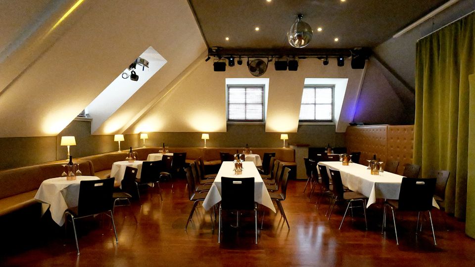 Gastraum unter einem schrägen Dach mit Dachfenstern, warmem Licht und Holzboden, ausgestattet mit Tischen mit weißen Tischdecken und schwarzen Stühlen. Entlang der Wände stehen gepolsterte Sitzbänke mit kleinen Tischlampen; an der Decke sind Spotlights und eine Discokugel angebracht.